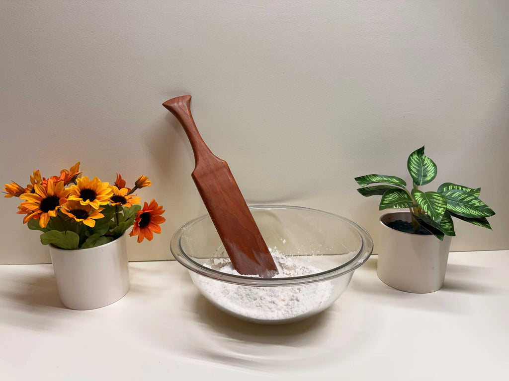 Wooden kitchen spurtle resting in a clear bowl of flour, reddish-brown finish.