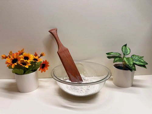 Wooden kitchen spurtle resting in a clear bowl of flour, reddish-brown finish.